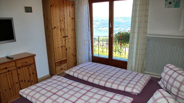 Ein Schlafzimmer mit zwei Betten, einem Holzschrank, einem Fernseher und einem Fenster mit Blick auf Grünflächen und Hügel.