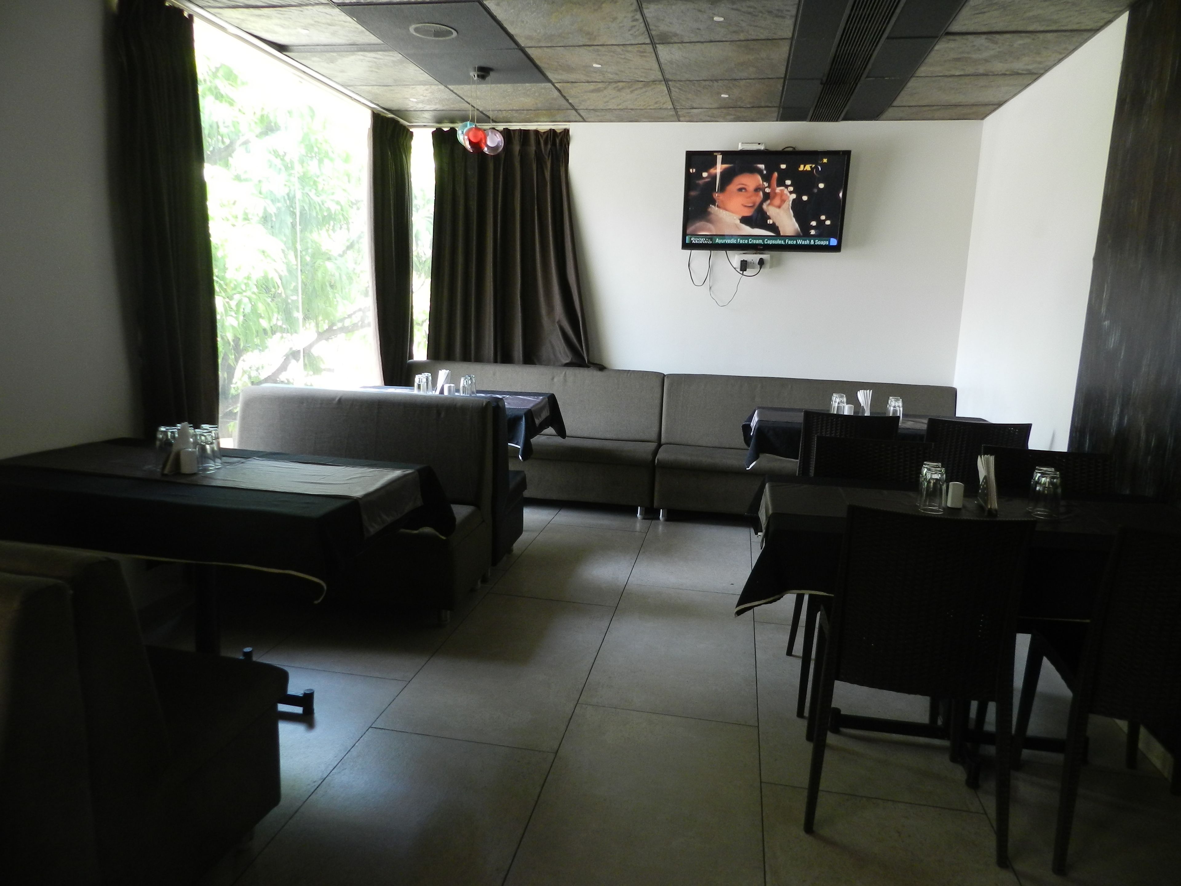A modern dining area with a sofa, tables, and chairs, a television mounted on the wall, and a view of greenery outside.