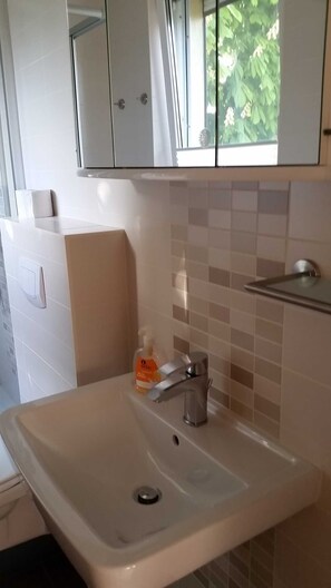 Un baño con un lavabo blanco, un espejo encima y una ventana con follaje en el exterior.