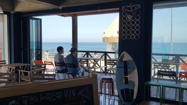 An indoor seating area with a view of the sea, a surfboard, and a chalkboard with text. - Donghae Sol Guesthouse - Hostel (Donghae)