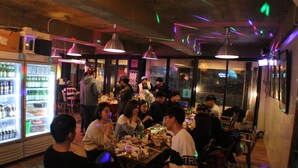 A crowded indoor dining area with tables and chairs, a refrigerator stocked with beverages, and hanging pendant lights. - Donghae Sol Guesthouse - Hostel (Donghae)