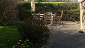 Un patio con suelo de piedra, sillas de madera, una mesa y un muro de piedra con plantas.