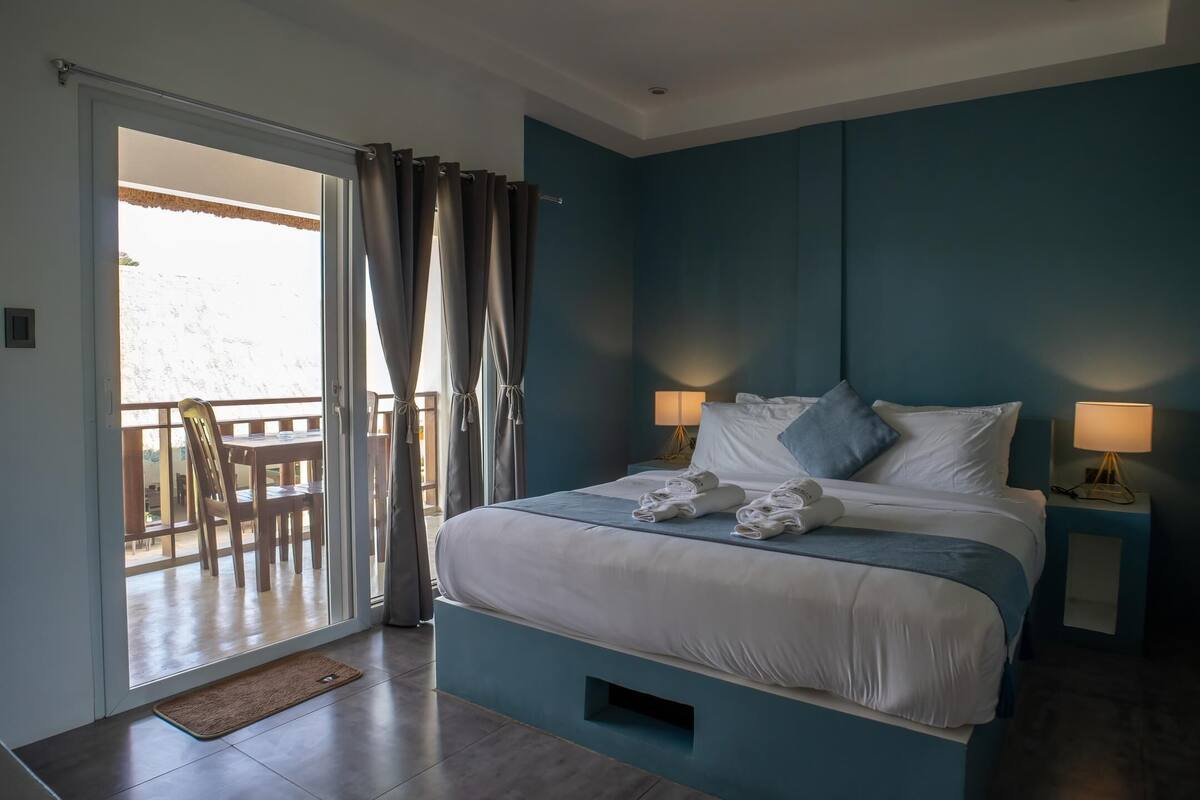 A bedroom with a bed, two bedside tables with lamps, a balcony with a chair, and a view of the outdoors.