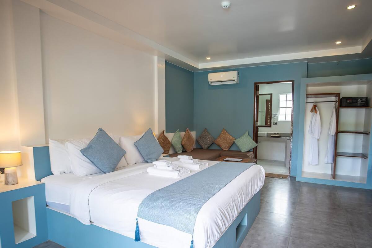 A modern hotel room with a large bed, a bedside table with a lamp, a bathroom with a mirror and towel rack, and a wardrobe.