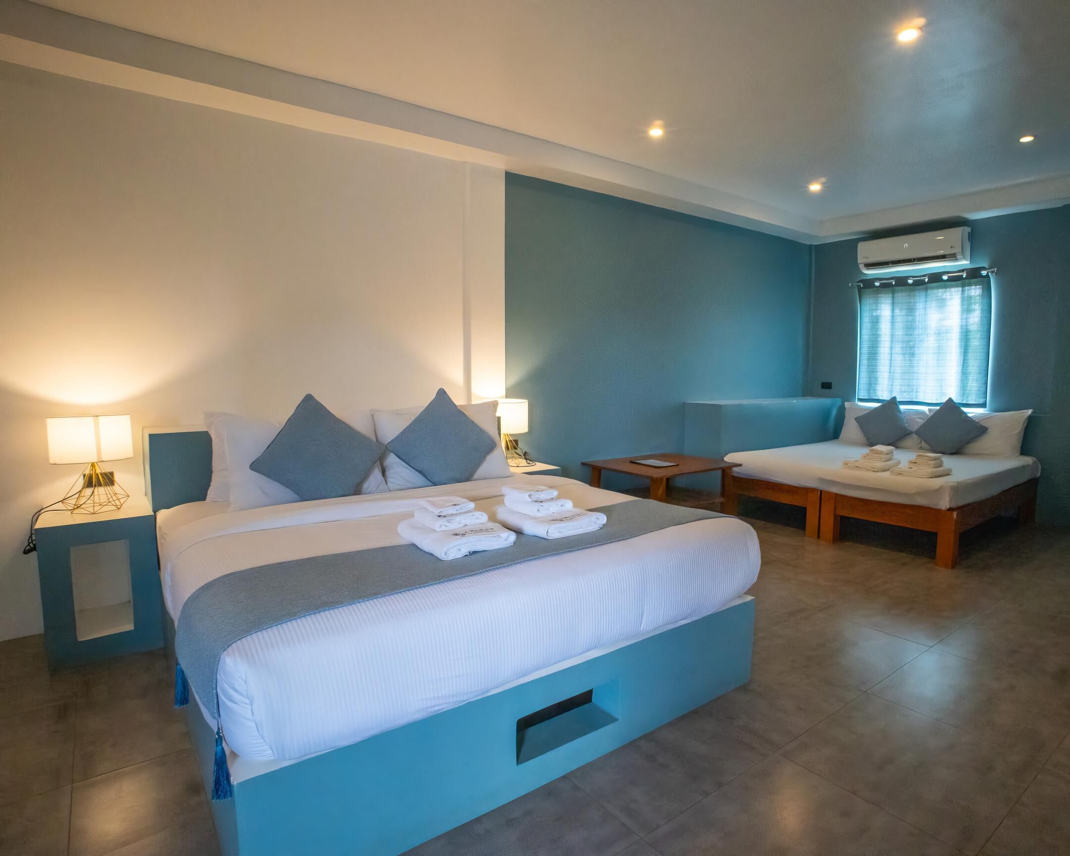 A hotel room with a large bed, a smaller sofa, a nightstand with a lamp, and an air conditioning unit.