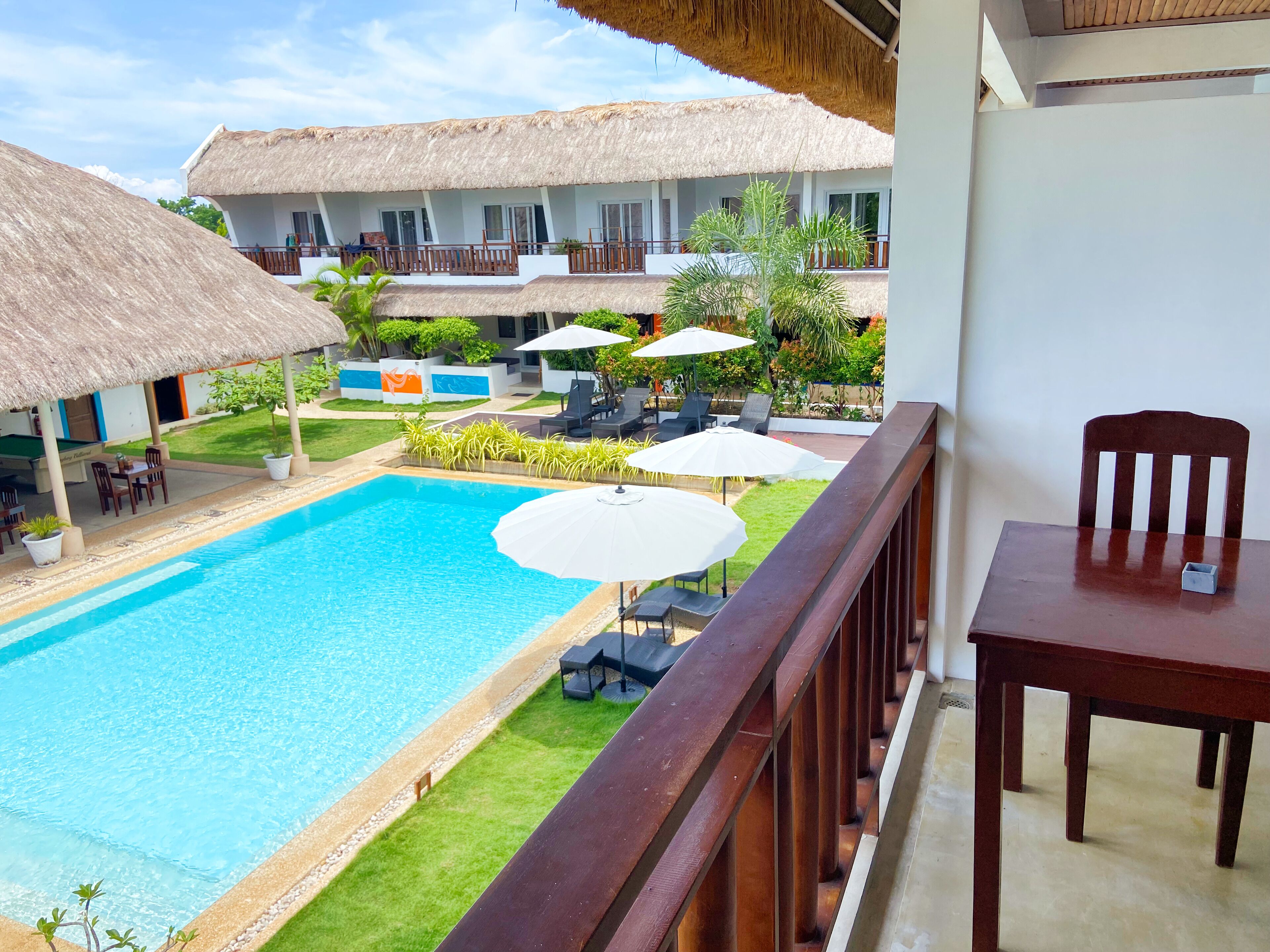 A poolside view with a wooden deck, lounge chairs, umbrellas, and a thatched-roof building.