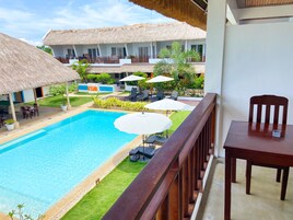 A poolside view with a wooden deck, lounge chairs, umbrellas, and a thatched-roof building.