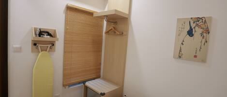 A compact bedroom with a bed, a small shelf with a kettle, a hanging clothes rack, and a window with blinds.