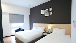 A hotel room with two beds, a large window with blinds, a dark wall with framed pictures, and a bedside table with a lamp. - De House Hotel (Sibu)