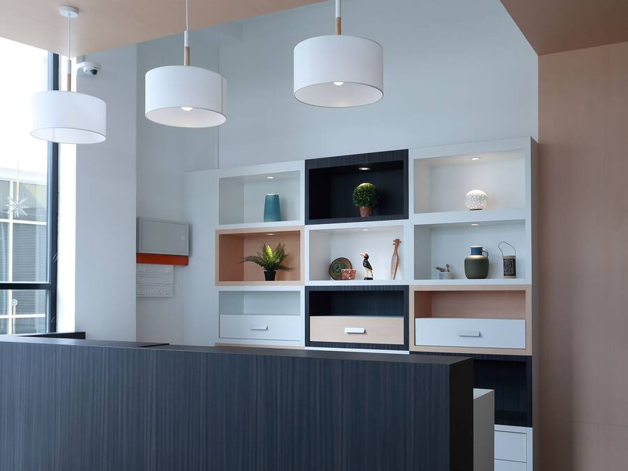 A modern reception desk with a grey panel, white shelving with various decorative items, and three pendant lights.