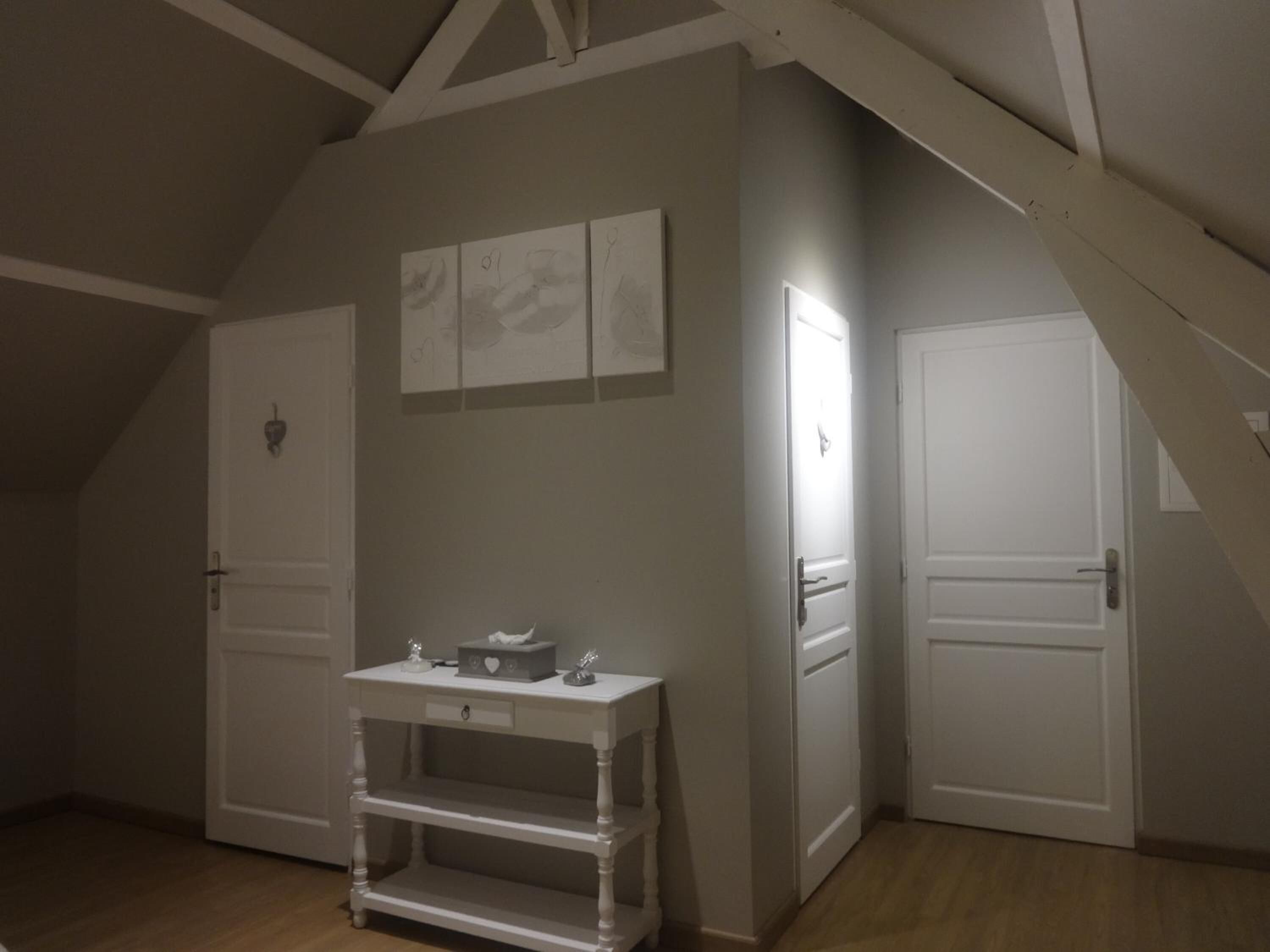 Una habitación con techo inclinado, una puerta blanca, una mesa blanca con cajones y un estante fijado a la pared.