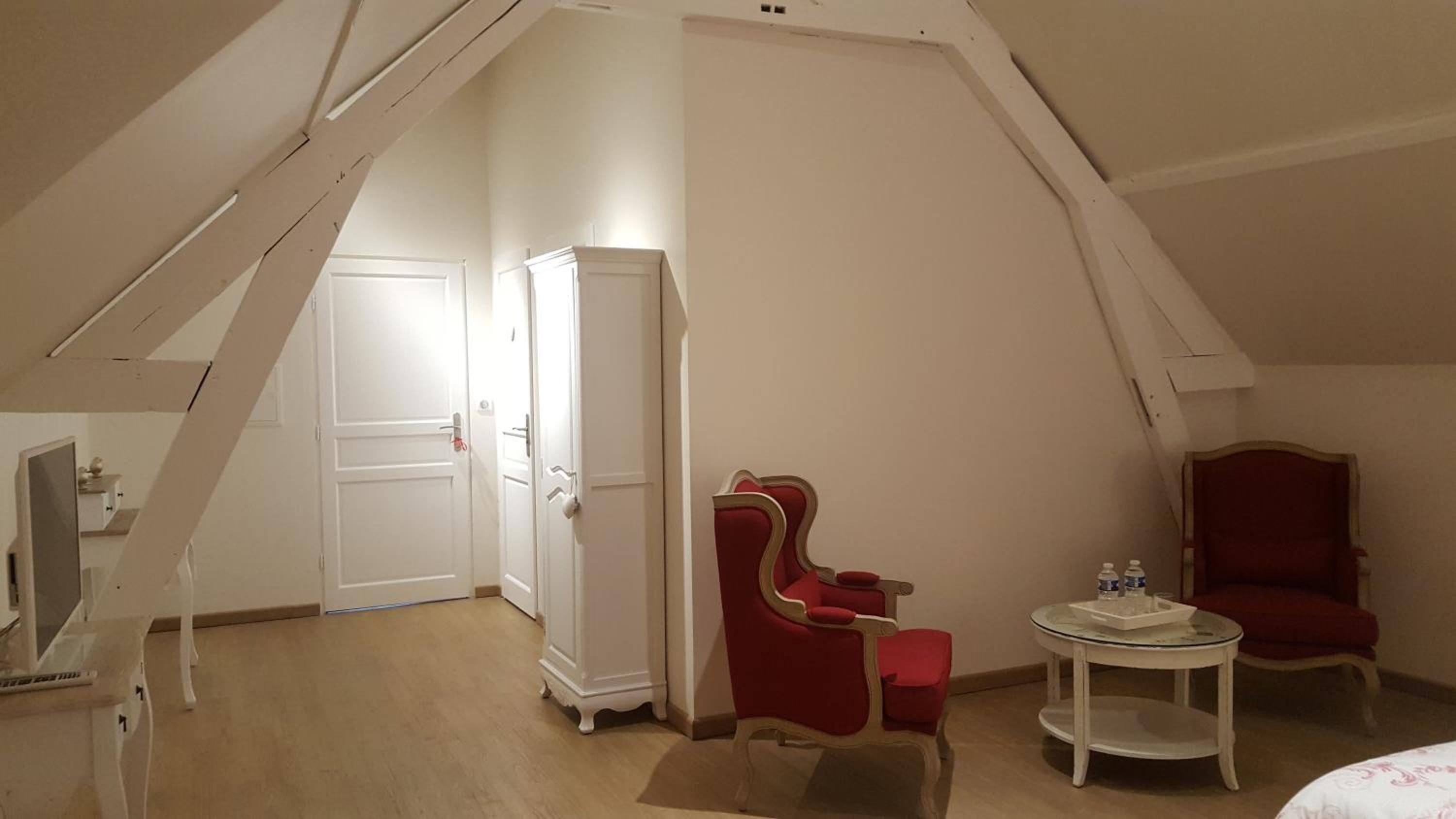A small, cozy room with a sloped ceiling, a red armchair, a matching chair, a small round table, and a wardrobe.