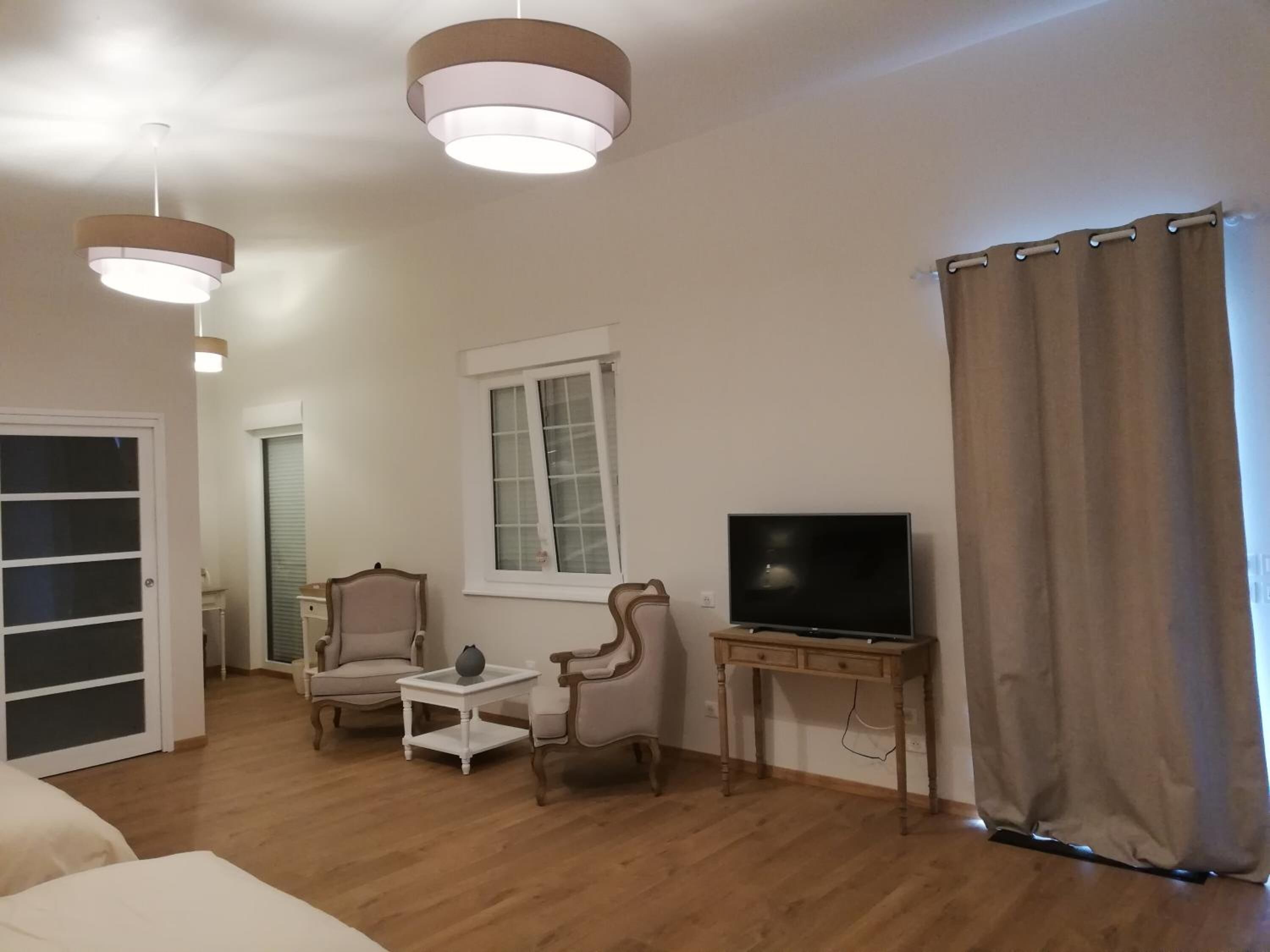 Un salon doté d’un parquet, d’un téléviseur posé sur un meuble en bois, de deux fauteuils, d’une petite table blanche, d’une fenêtre avec des rideaux et d’une porte avec un panneau de verre.