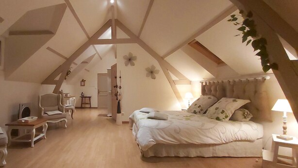 Un acogedor dormitorio en el ático con techo inclinado, cama con ropa de cama floral, una silla, una mesita y una lámpara.