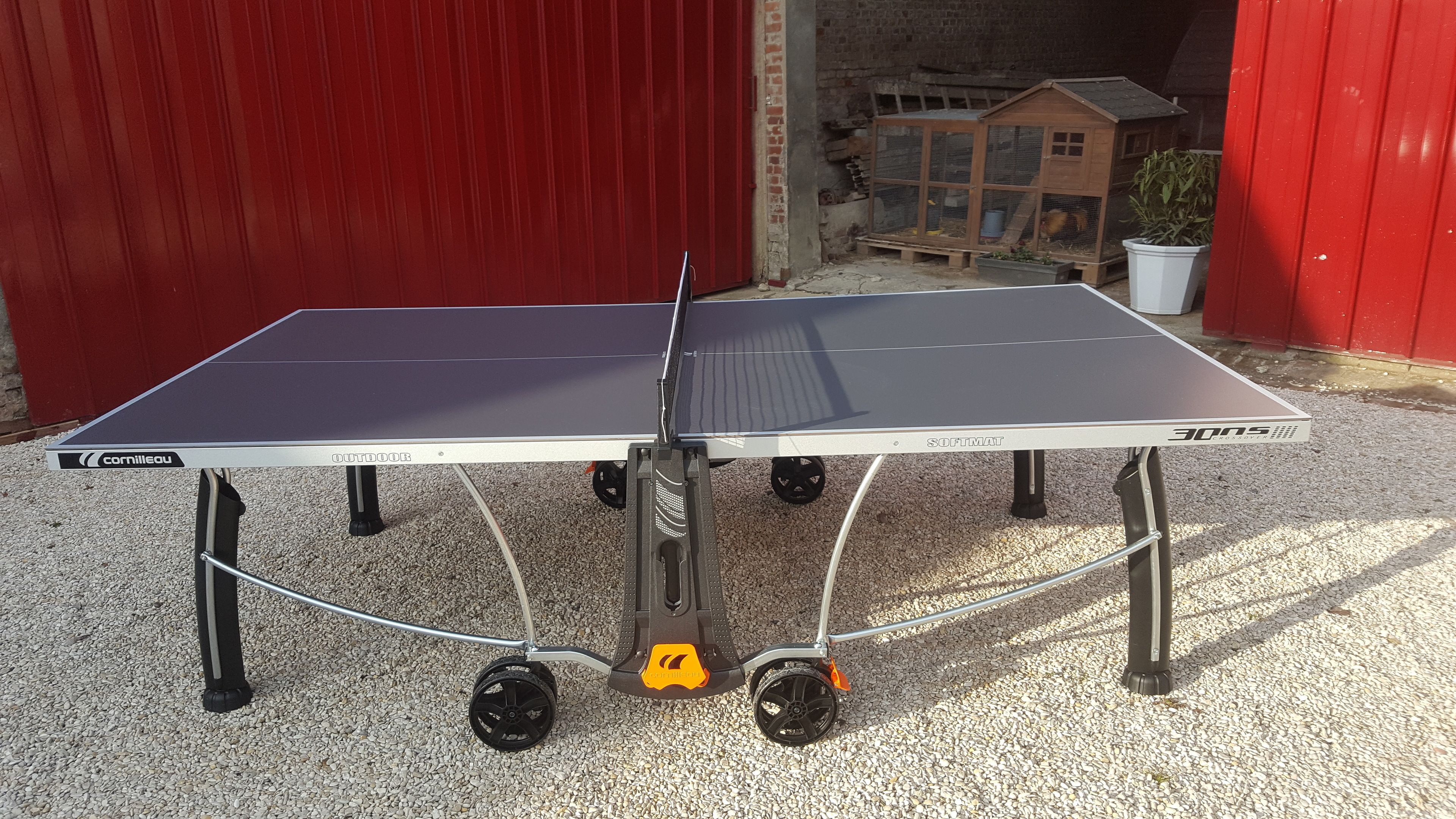Una mesa de ping pong portátil con ruedas, etiquetada 'Cornilleau OUTDOOR' y 'SOFTMAT'.