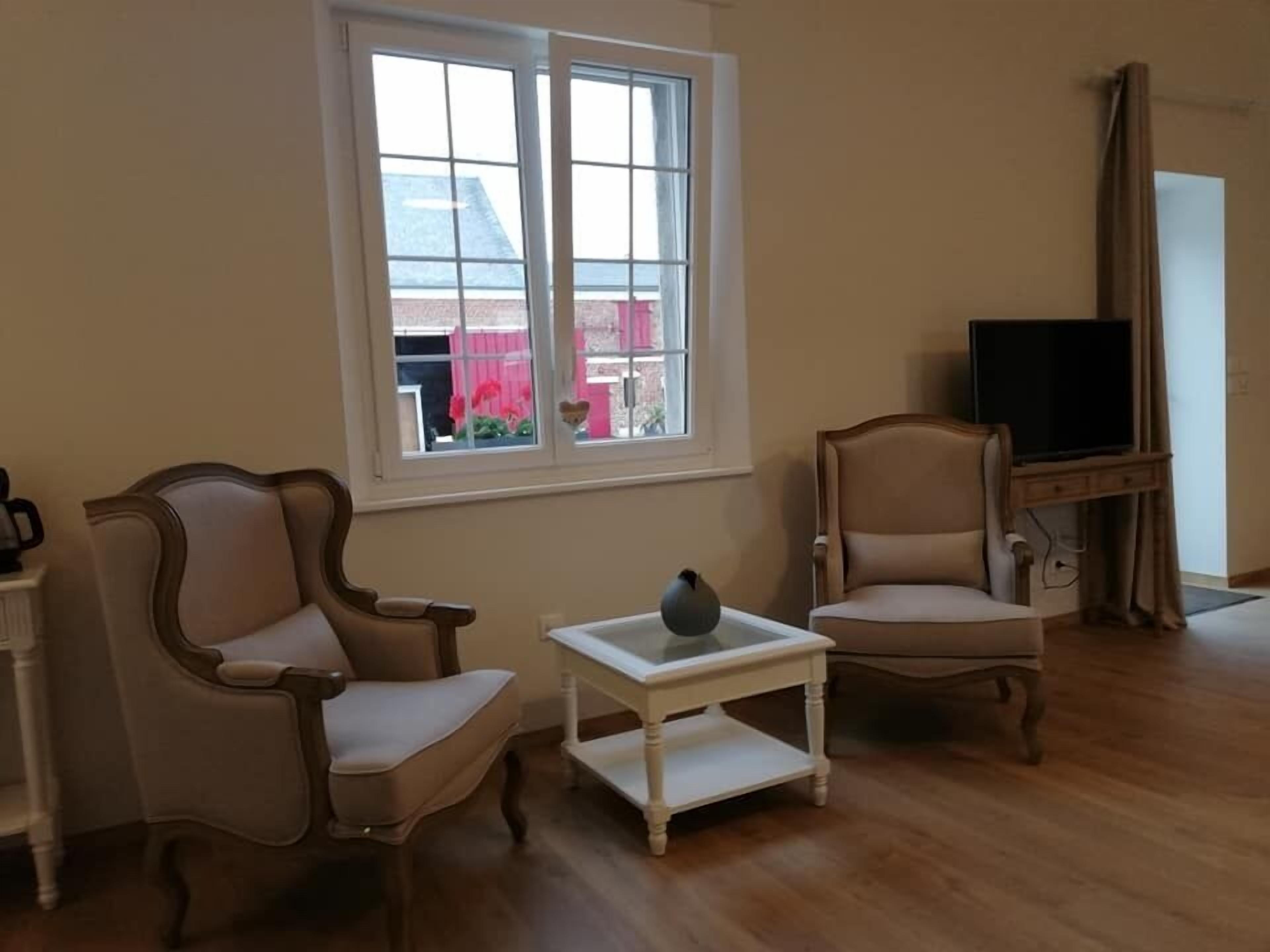 A living room with two armchairs, a small white table, a television on a stand, and a window with a view of buildings.