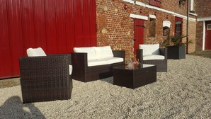 Un espacio al aire libre con mobiliario de mimbre y una mesa con cubierta de vidrio, frente a una pared roja y un edificio de ladrillo.