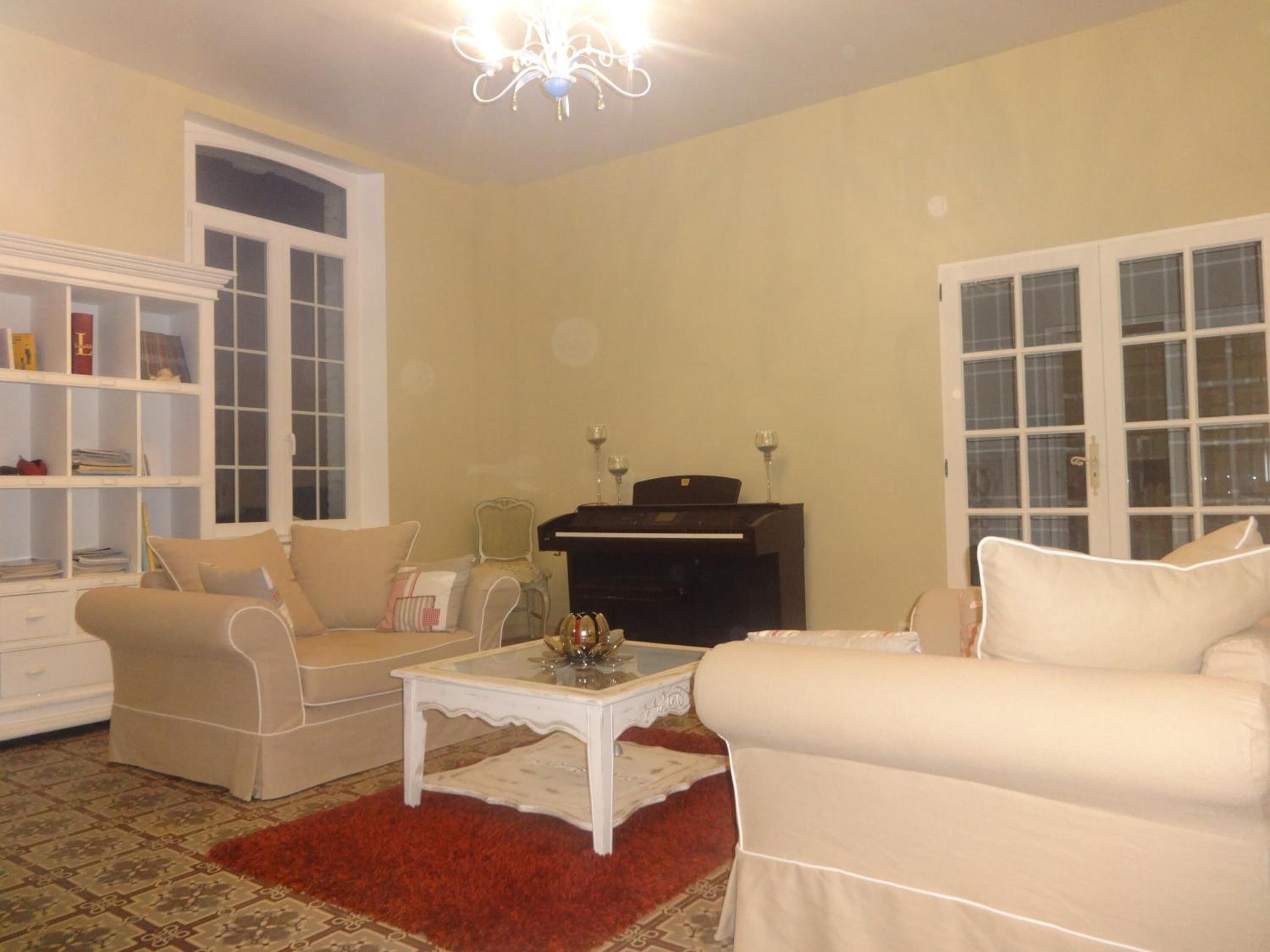 A living room with a beige sofa, a white armchair, a glass coffee table, a bookshelf, a piano, and a red rug.
