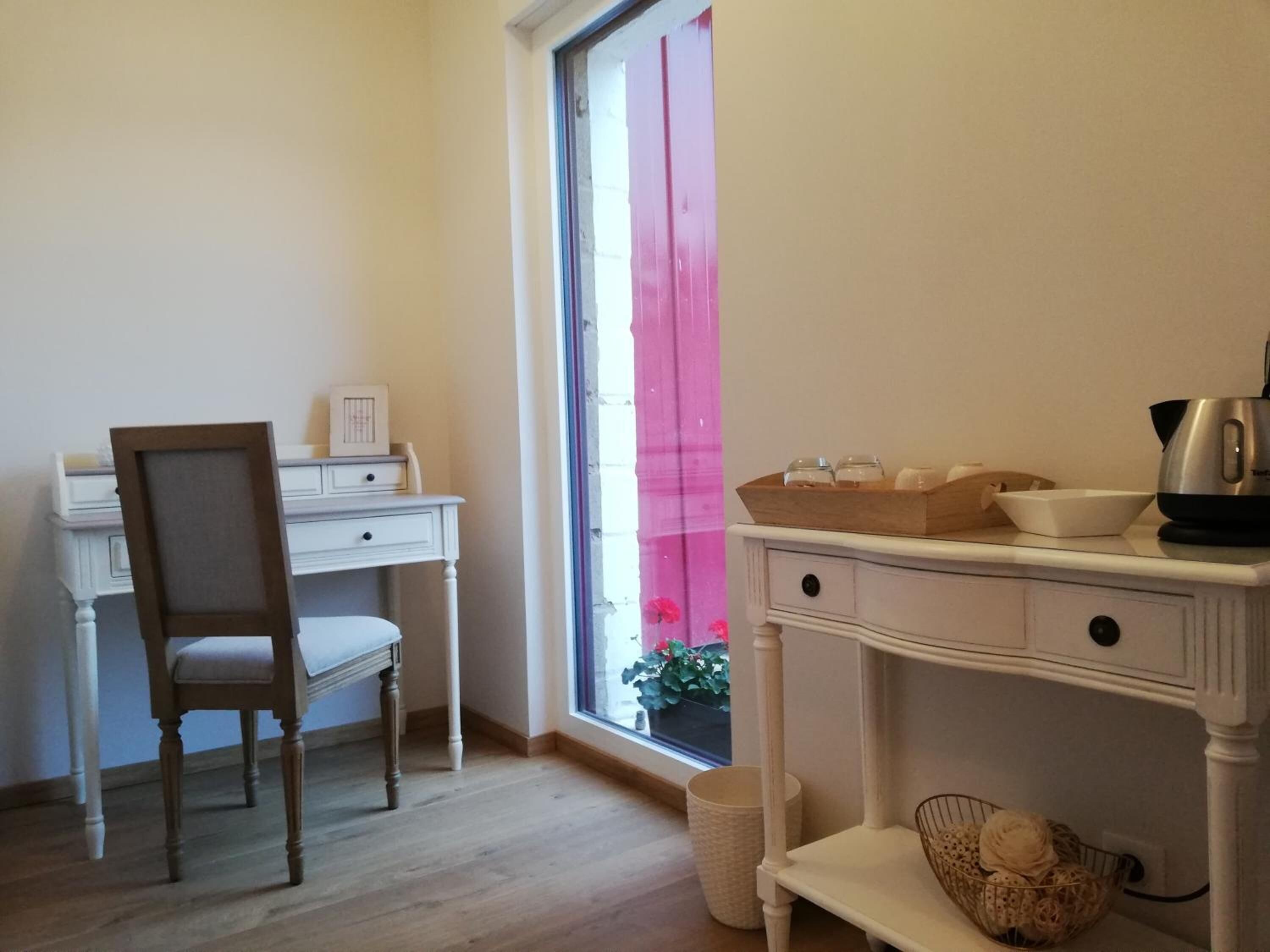 Een kamer met een houten bureau, een stoel, een raam met een roze gordijn en een witte console met keukenbenodigdheden.