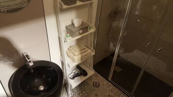 A bathroom with a black sink, a toilet, a glass shower enclosure, and a shelf with towels and toiletries.