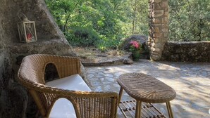 A wicker chair and stool set on a stone patio with a view of a lush garden. - small, cozy eco-house for environmentally conscious nature-lover (Saint-Roman-De-Codières)