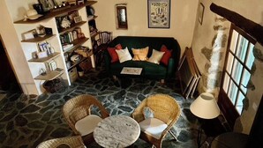 A cozy room with a stone floor, a green sofa with cushions, wicker chairs, a marble table, and a bookshelf filled with books and decorative items. - small, cozy eco-house for environmentally conscious nature-lover (Saint-Roman-De-Codières)