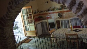 A rustic stone-walled room with a kitchen area, including a stove, refrigerator, and a dining table with chairs. - small, cozy eco-house for environmentally conscious nature-lover (Saint-Roman-De-Codières)