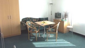 A small, sparsely furnished room with a single table and two chairs, a sofa, a cabinet, and a television.