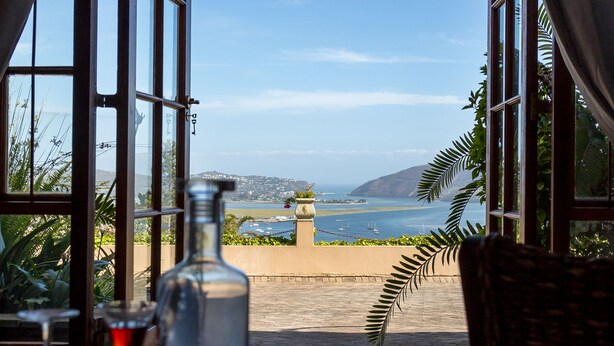 A table with a bottle and a glass, a wicker chair, and a view of the sea through large windows. - Moncrieff Manor (Knysna)