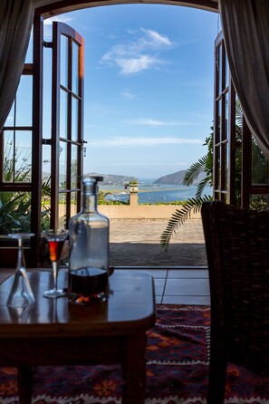 Una mesa con una botella y un vaso, una silla de mimbre y vista al mar a través de amplios ventanales.