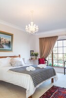 Un dormitorio con una cama grande, una araña, una ventana con cortinas, un cuadro y una pequeña mesa con un jarrón.
