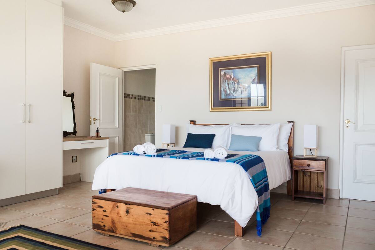 A bedroom with a bed, bedside tables, a wooden chest, a painting on the wall, and a bathroom door.