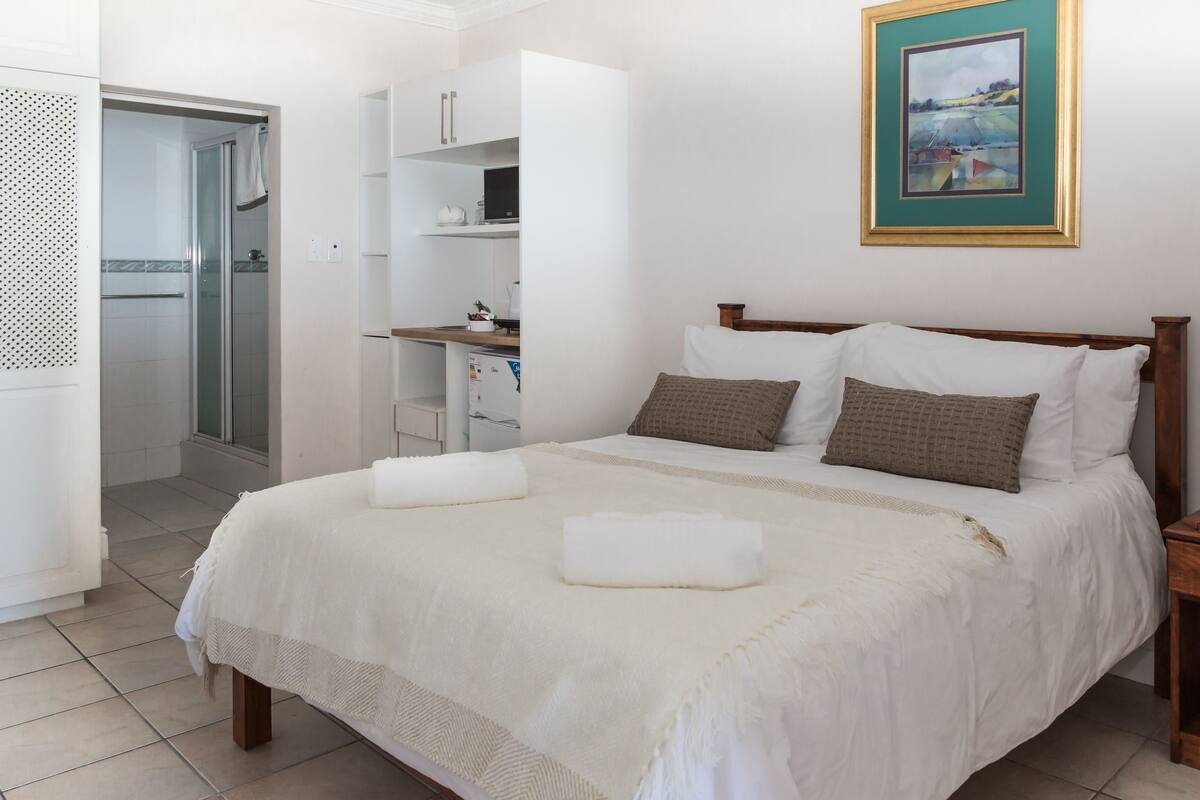 A neatly made bed with a white comforter and two pillows, a wooden nightstand with a lamp, and a framed picture on the wall.