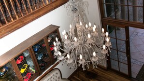 A room with a chandelier, a bench, a display of model cars, and a large window with a view of trees and buildings. - Moncrieff Manor (Knysna)