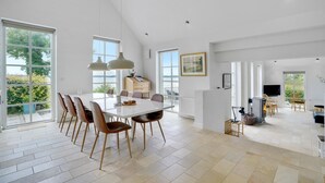 Un comedor amplio con una mesa y sillas blancas, ventanas grandes con vistas a zonas verdes, y una zona de cocina con televisor y sillas de mimbre.
