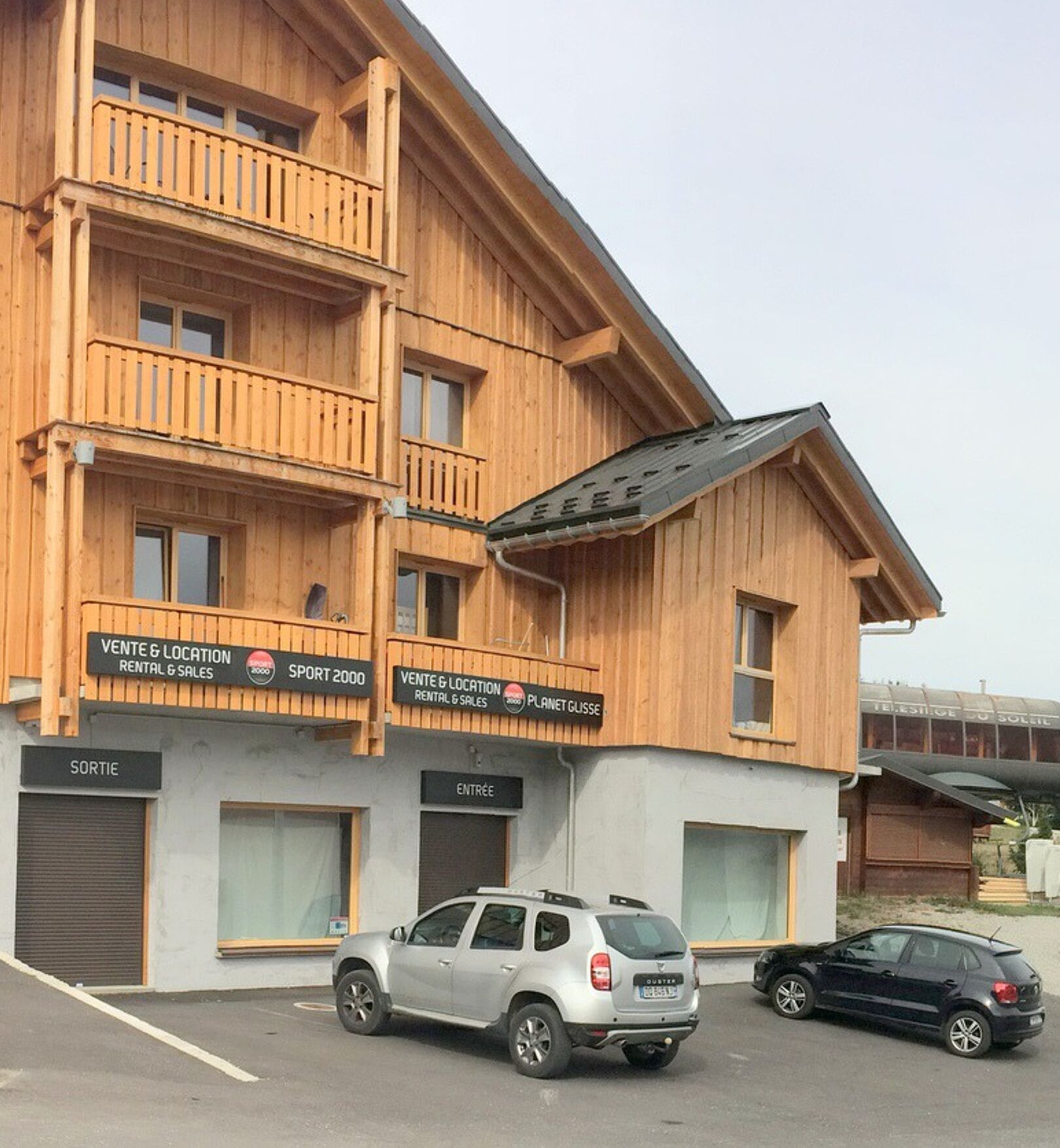 Un edificio de madera de varios pisos con balcones, carteles que dicen 'VENTE & LOCATION RENTAL & SALES' y 'SPORT 2000', y un estacionamiento con dos coches.
