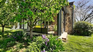 A modern house with a stone exterior, a covered porch with outdoor seating, and a lush garden with purple flowers. - Elegant stone house in the countryside with swimming pool (Pressignac-Vicq)