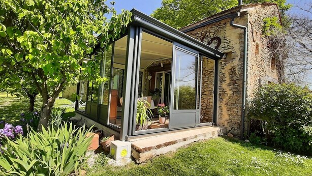 A glass-enclosed room with a stone exterior, surrounded by lush greenery and flowering plants. - Elegant stone house in the countryside with swimming pool (Pressignac-Vicq)
