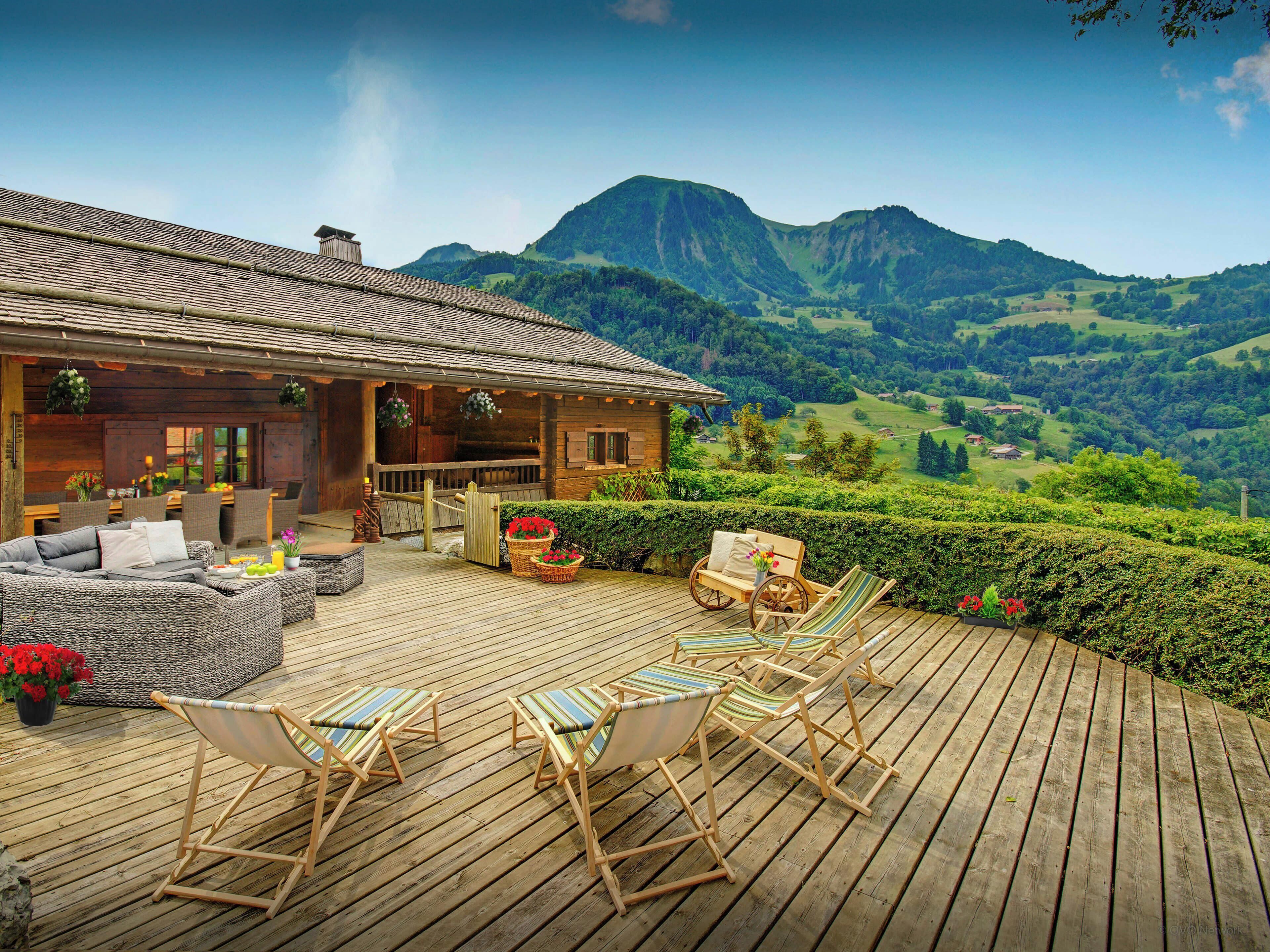 Eine Holzterrasse mit Gartenmöbeln, ein Holgebäude und eine gebirgige Landschaft im Hintergrund.