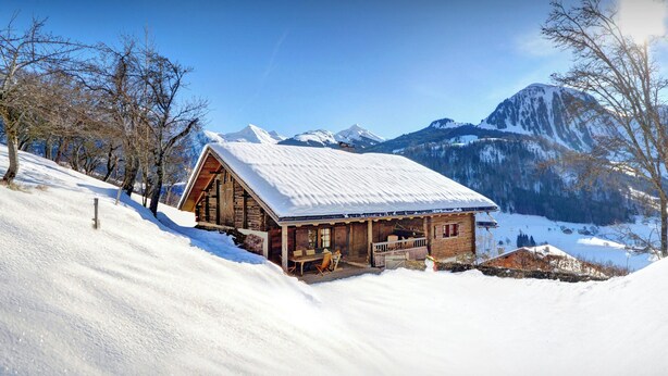 Eine Holzhütte mit schneebedecktem Dach, umgeben von schneebedeckten Bergen und Bäumen.