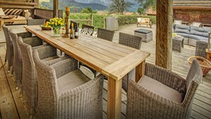 Eine Holzterrasse mit Korbmöbeln, einem Tisch mit Weingläsern und Flaschen sowie einem Panoramablick auf die Berge.