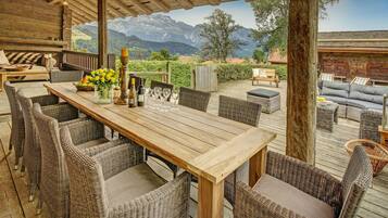 A wooden deck with wicker furniture, a table set with wine glasses and bottles, and a scenic mountain view.