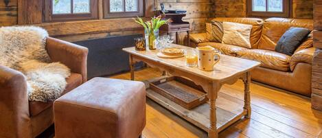 A cozy log cabin interior with a leather sofa, a fluffy chair, a wooden coffee table, and a fireplace.