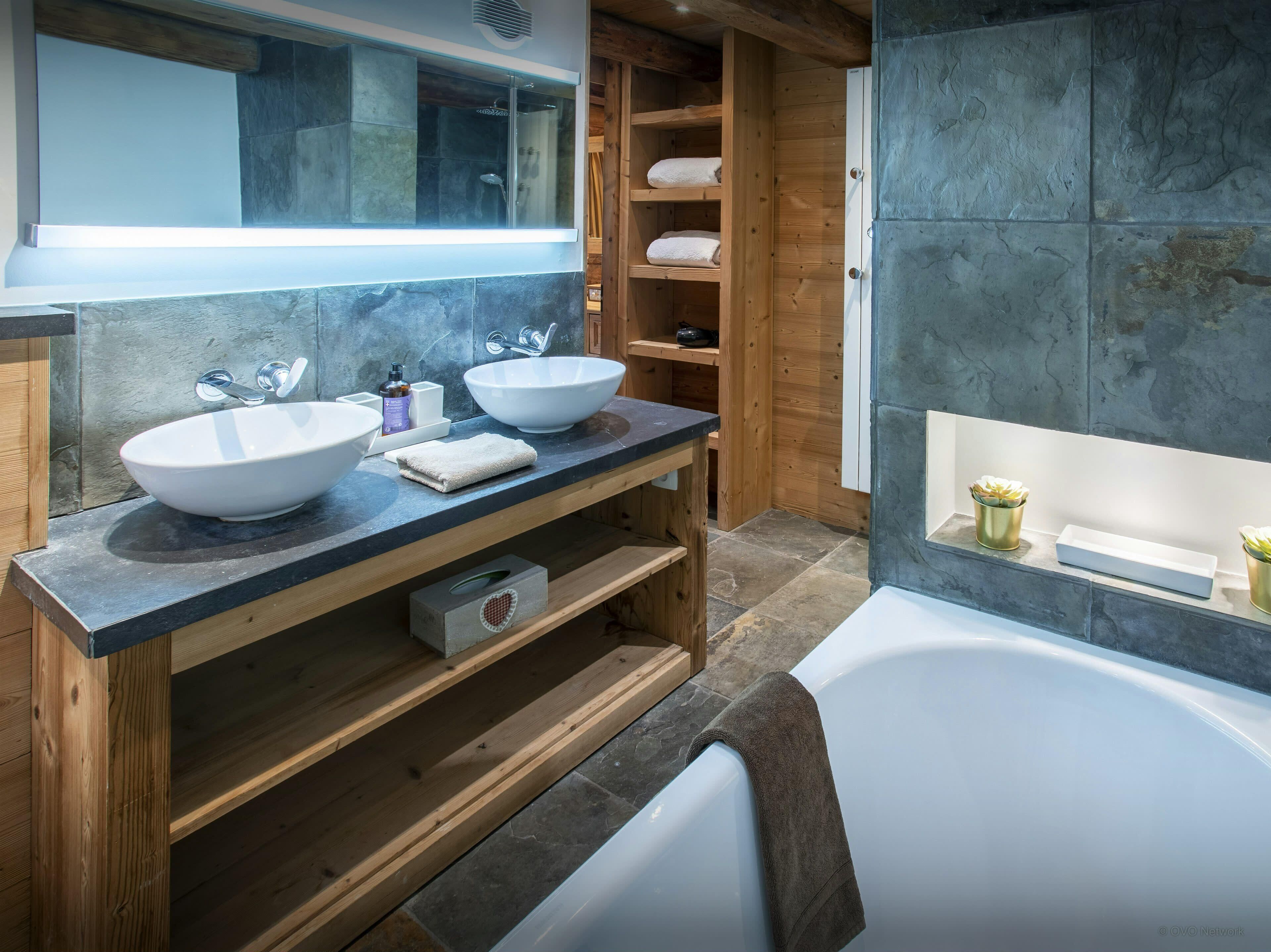 Une salle de bain moderne avec deux grands lavabos, une baignoire, des étagères en bois et un mur en pierre.