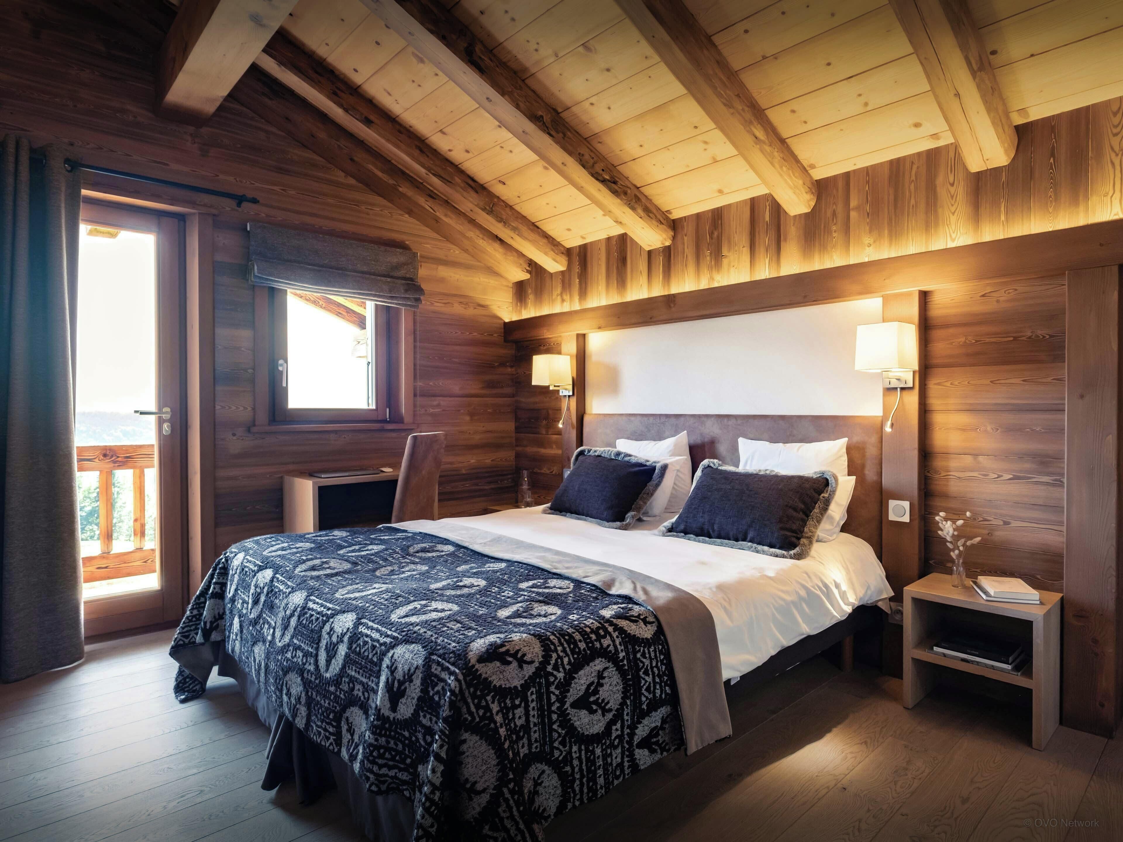 A wooden cabin bedroom with a bed, bedside lamps, and a view through a window.