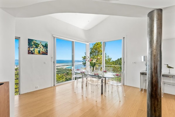 A modern dining area with a glass table, clear chairs, and a view of the ocean.