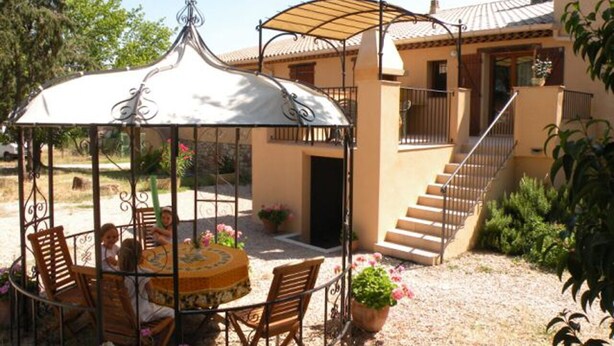 Un área de comedor al aire libre con techo, que incluye una mesa y sillas, rodeada de plantas en macetas y una escalera que sube a un nivel superior.