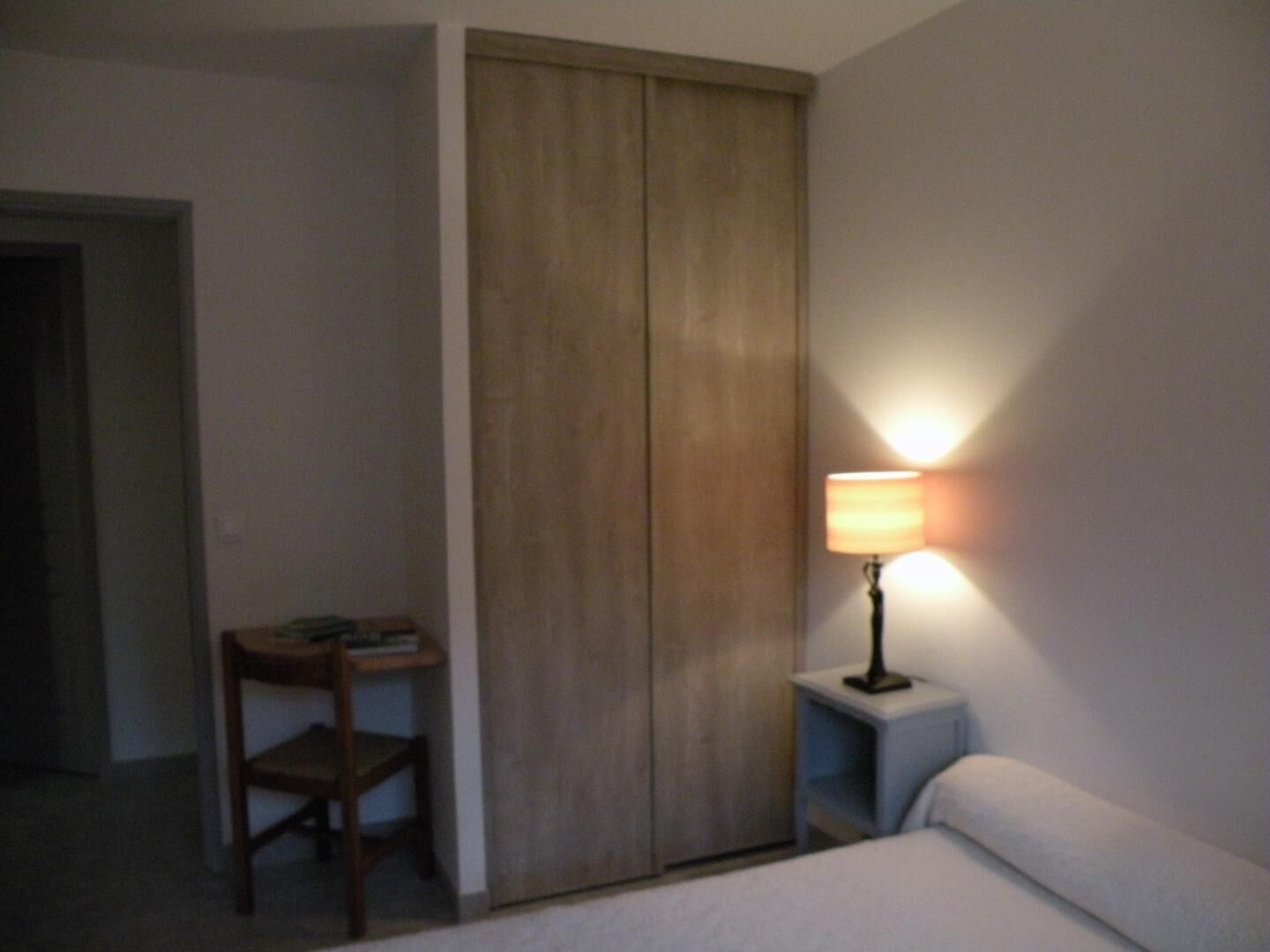 A hotel room with a bed, a desk with a chair, a wardrobe, and a bedside table with a lamp.