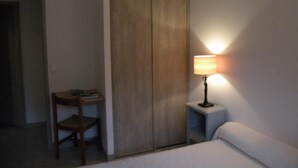 Habitación de hotel con cama, escritorio con silla, armario y mesita de noche con lámpara.