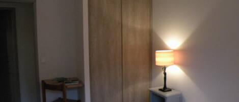 A hotel room with a bed, a desk with a chair, a wardrobe, and a bedside table with a lamp.